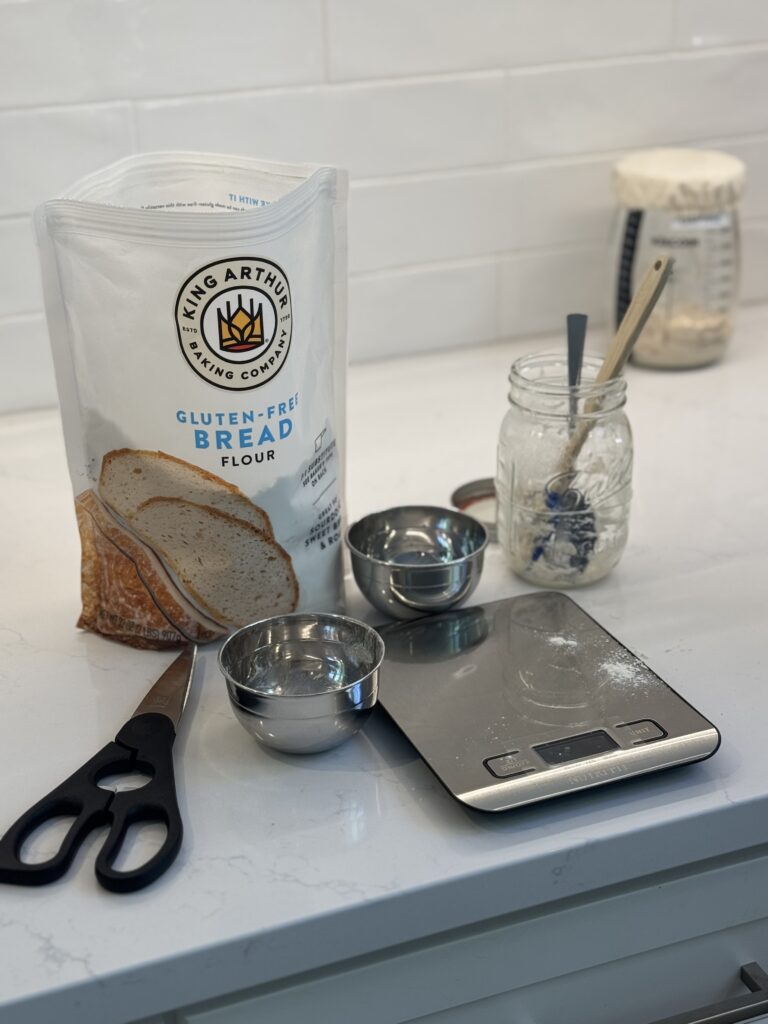 King Arthur Gluten Free bread flour with kitchen scale measuring cups and sourdough starter on a white quartz counter